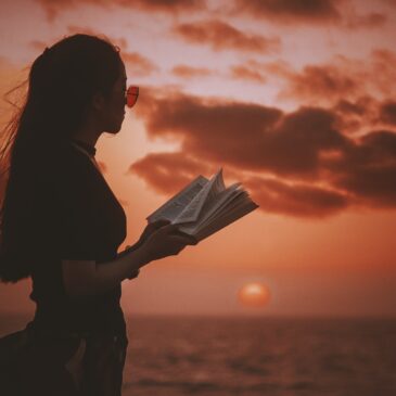 woman holding book while looking at body of water during golden hour